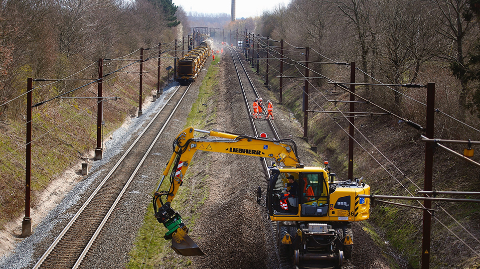 Gravemaskine i gang med sporbyggeri for Banedanmark mens der kører tog modsat i det andet spor